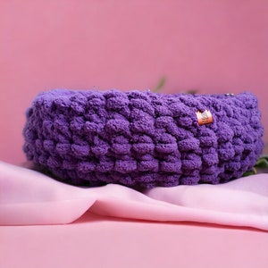 May include: Purple crocheted bowl with a small brown label that says "Hand Made". The bowl is sitting on a pink fabric surface.