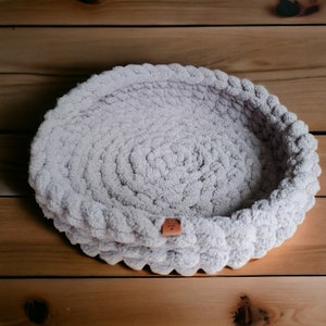 May include: A light gray crocheted pet bed with a brown leather tag. The bed is round and has a soft, fluffy texture.