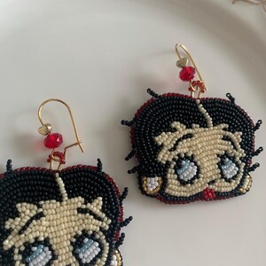 Pode incluir: Um par de brincos de contas com um personagem de desenho animado com cabelo preto, pele branca e olhos azuis. Os brincos s&atilde;o feitos com contas pretas, brancas, vermelhas e douradas.