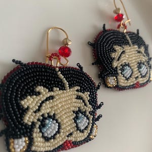 Pode incluir: Um par de brincos de contas com um personagem de desenho animado com cabelo preto, pele branca e olhos azuis. Os brincos s&atilde;o feitos com contas pretas, brancas, vermelhas e douradas.