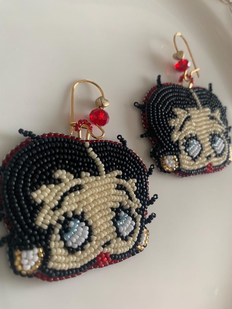 Pode incluir: Um par de brincos de contas com um personagem de desenho animado com cabelo preto, pele branca e olhos azuis. Os brincos s&atilde;o feitos com contas pretas, brancas, vermelhas e douradas.