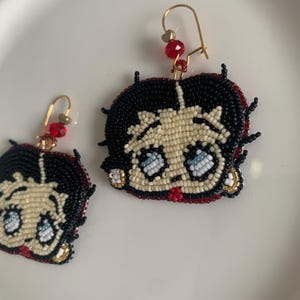 Pode incluir: Um par de brincos de contas com um personagem de desenho animado com cabelo preto, pele branca e olhos azuis. Os brincos s&atilde;o feitos com contas vermelhas, pretas, brancas e douradas.