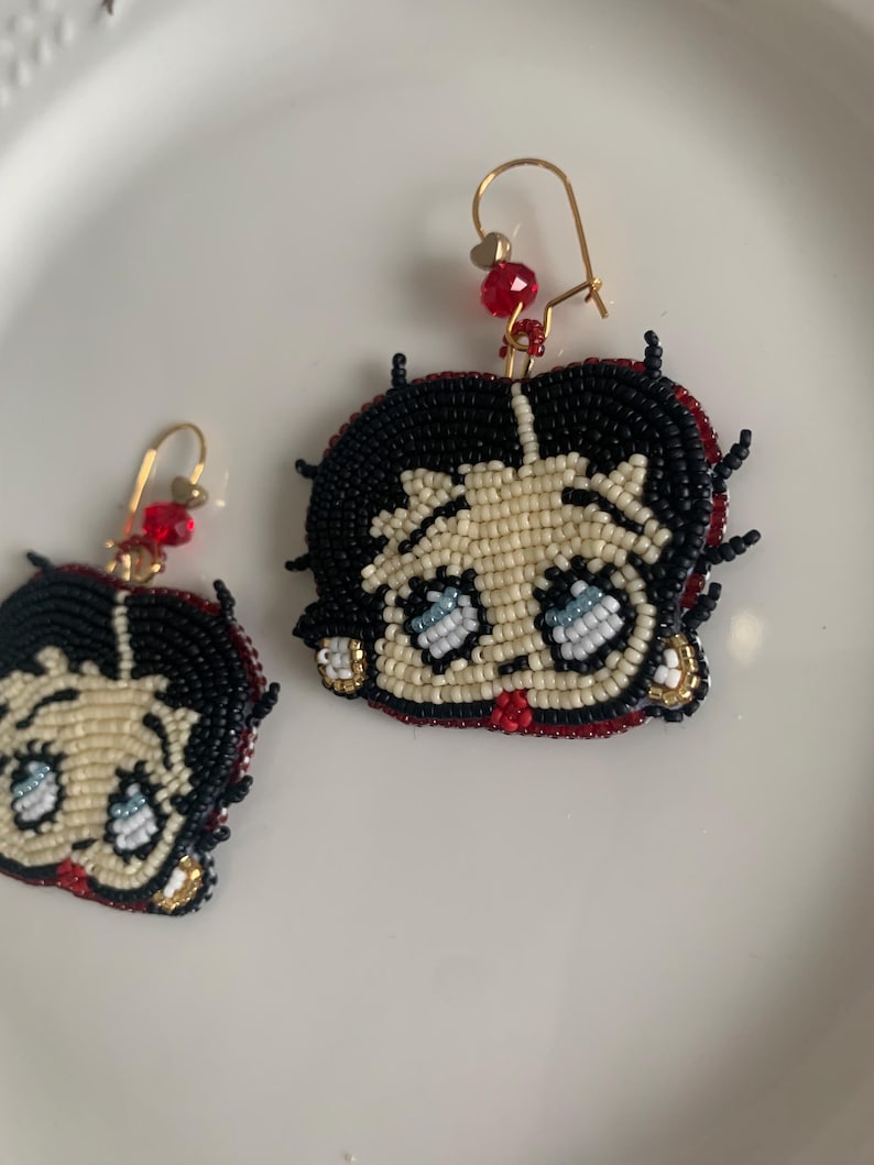 Pode incluir: Um par de brincos de contas com um personagem de desenho animado com cabelo preto, pele branca e olhos azuis. Os brincos s&atilde;o feitos com contas vermelhas, pretas, brancas e douradas.