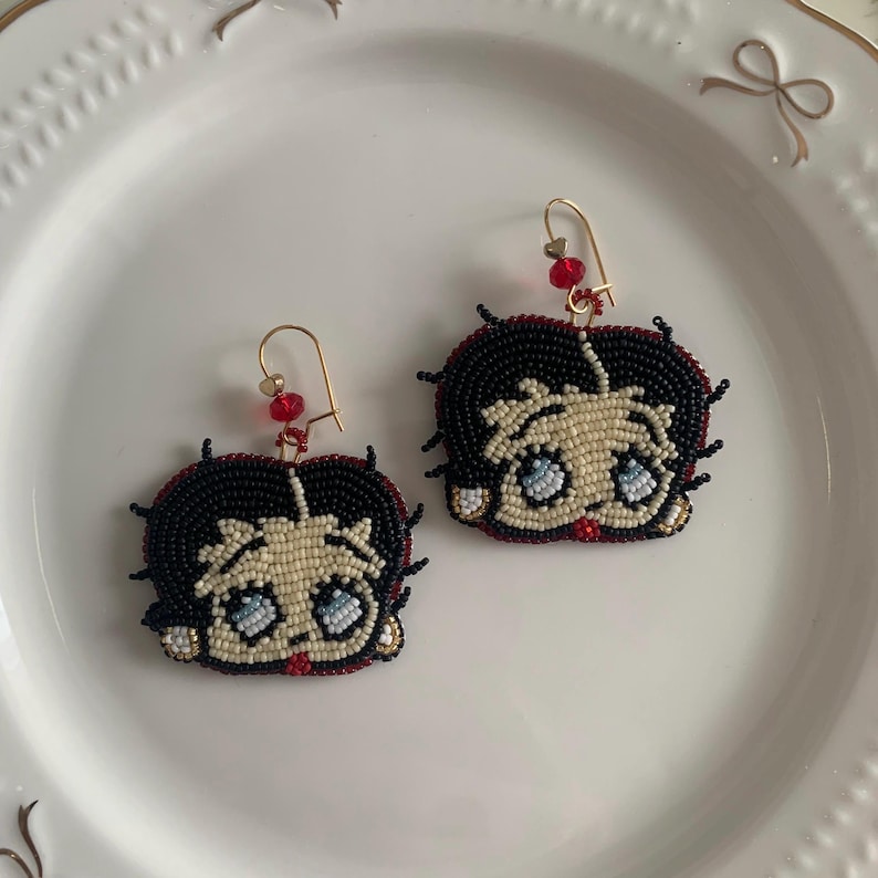 Pode incluir: Um par de brincos de contas com um personagem de desenho animado com cabelo preto, pele branca e olhos azuis. Os brincos s&atilde;o feitos com contas pretas, brancas, vermelhas e azuis.