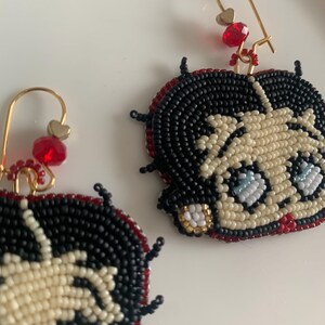 Pode incluir: Um par de brincos de contas com um personagem de desenho animado com cabelo preto, pele branca e olhos azuis. Os brincos s&atilde;o feitos com contas vermelhas, brancas, pretas e douradas. Os brincos s&atilde;o presos a ganchos dourados com contas vermelhas e brancas.