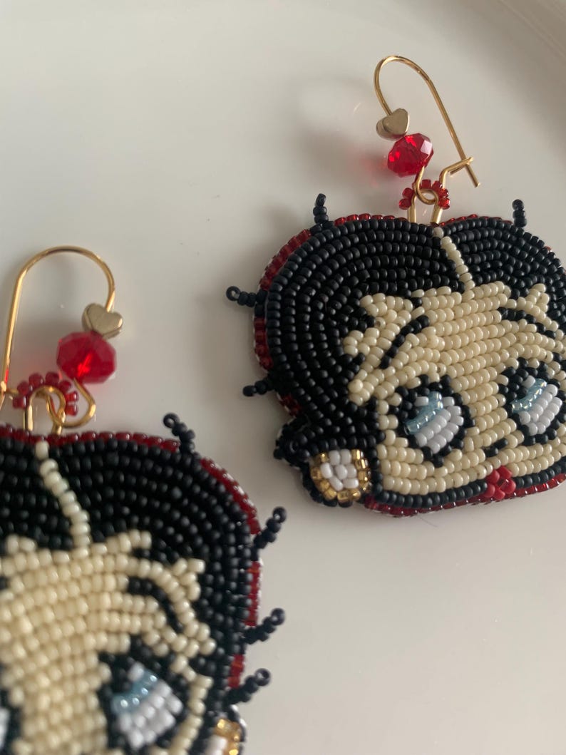 Pode incluir: Um par de brincos de contas com um personagem de desenho animado com cabelo preto, pele branca e olhos azuis. Os brincos s&atilde;o feitos com contas vermelhas, brancas, pretas e douradas. Os brincos s&atilde;o presos a ganchos dourados com contas vermelhas e brancas.