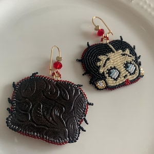 Pode incluir: Um par de brincos de contas, um com um personagem de desenho animado com cabelo preto e olhos azuis, e o outro com um design de cora&ccedil;&atilde;o de couro marrom.