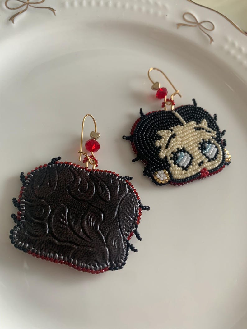 Pode incluir: Um par de brincos de contas, um com um personagem de desenho animado com cabelo preto e olhos azuis, e o outro com um design de cora&ccedil;&atilde;o de couro marrom.