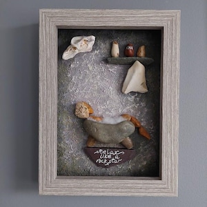 Puede incluir: Obra de arte enmarcada con guijarros que representa a una persona relajándose en una bañera de piedra. La obra presenta una combinación de colores gris y marrón, con el texto "relax like a rock star" en un letrero marrón. Otros guijarros están dispuestos encima de la bañera.