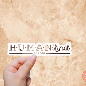 May include: White sticker with the text "H.U.M.A.N.Kind" in brown lettering with a line under the text. The text "BE BOTH" is below the line. The sticker is being held by a hand against a beige background.