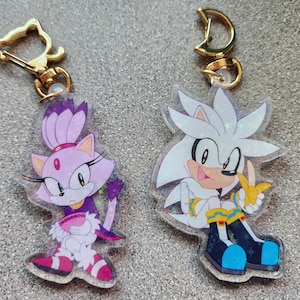 May include: Two character keychains featuring Sonic the Hedgehog characters. The keychain on the left is a pink and purple Blaze the Cat keychain. The keychain on the right is a silver and white Silver the Hedgehog keychain.
