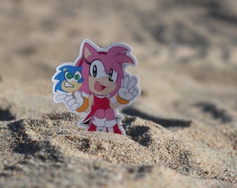 Puede incluir: Un personaje de dibujos animados rosa y blanco con ojos azules y un vestido rosa, sosteniendo un personaje de dibujos animados azul y blanco con una piruleta amarilla. Los personajes están en una playa de arena.