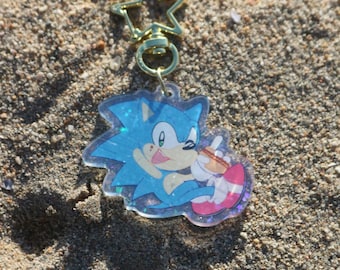 May include: A blue and pink glittery keychain featuring a cartoon character of Sonic the Hedgehog holding a hot dog. The keychain is attached to a gold star-shaped ring.