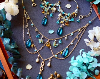 Può includere: Una collana dorata e orecchini a cerchio abbinati con gemme blu a forma di goccia, piccole perline e dettagli a farfalla. I gioielli sono esposti su una superficie grigio scuro, circondati da fiori secchi in un vassoio di legno.