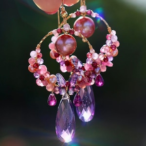 May include: Elegant gold hoop earrings adorned with pink and purple gemstones and pearls. The earrings feature large, faceted teardrop-shaped purple gems and smaller, round pink and purple beads, creating a delicate and luxurious design.