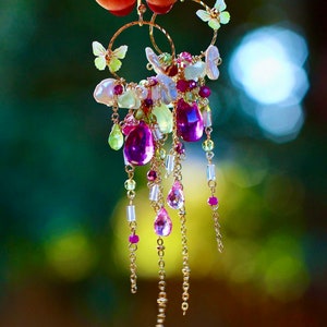 May include: Gold hoop earrings with dangling purple, pink, and green gemstones. The earrings feature small butterfly accents and pearl-like beads. The design includes delicate chains and a variety of bead shapes, creating a whimsical and elegant look.