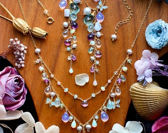 Può includere: Una collezione di gioielli color oro, tra cui una collana e orecchini, con varie pietre preziose e perle. La collana ha più fili con pendenti a forma di goccia. Gli orecchini hanno un design a cerchio con gemme pendenti. I gioielli sono esposti su una superficie di legno.