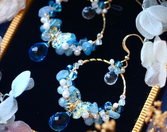 May include: A pair of elegant gold-toned hoop earrings. Each earring features a crescent design adorned with blue and clear gemstones, small white beads, and a large teardrop-shaped blue crystal. The earrings are set against a dark blue background.