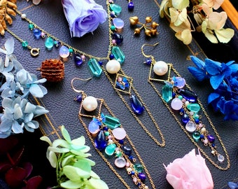May include: A gold-toned jewelry set featuring a necklace and earrings, adorned with blue, green, and pink gemstones, and white pearls. The jewelry is displayed on a dark surface, surrounded by dried flowers.