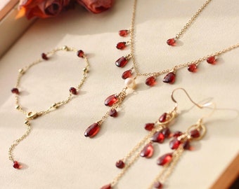 May include: A gold-toned jewelry set featuring a necklace, bracelet, and earrings. The necklace and earrings have dangling, teardrop-shaped red gemstones. The bracelet has small red gemstones and gold beads. A few red roses are in the background.