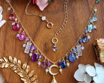 Può includere: Una delicata collana d'oro con un ciondolo a forma di mezzaluna e una grande gemma a forma di goccia. La collana presenta una varietà di pietre preziose colorate nelle tonalità del rosa, del viola e del blu. La collana è esposta su una superficie di legno.