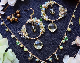 Può includere: Un set di gioielli color oro con collana e orecchini a cerchio. La collana e gli orecchini sono adornati con pietre preziose chiare, gialle e verdi, insieme a piccole perle bianche. I gioielli sono esposti su una superficie blu scuro, circondati da fiori bianchi e verde.