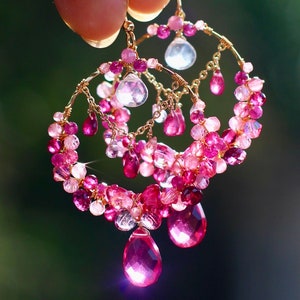 May include: Gold wire earrings with pink and fuchsia gemstones. The earrings feature a circular design with dangling teardrop-shaped gems and clusters of smaller beads in varying shades of pink.