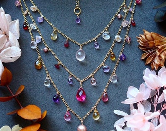 Può includere: Una collana a più fili con catene dorate e una varietà di pietre preziose colorate. La collana presenta pietre a goccia rosa, viola, gialle e trasparenti, insieme a un grande pendente di pietra rosa. Presentata su una superficie grigio scuro con accenti floreali.
