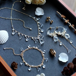 May include: A gold-toned jewelry set featuring a necklace, bracelet, and earrings. Each piece is adorned with clear, teardrop-shaped crystals and small white pearls. The jewelry is displayed on a dark gray surface with floral accents.