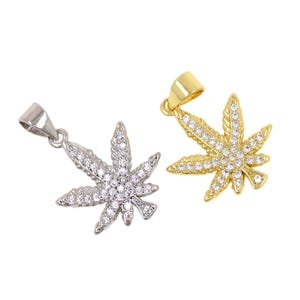 Puede incluir: Dos colgantes con forma de hoja de cannabis. Uno es plateado, el otro dorado. Ambos están incrustados con pequeños cristales transparentes. Los colgantes se muestran sobre un fondo blanco.