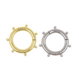 May include: Two round metal clasps, one gold-toned and one silver-toned, are displayed against a white background. Each clasp has a circular design with small, evenly spaced decorative elements around the outer edge.