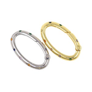 Peut inclure: Deux fermoirs ovales, un argenté et un doré, sont présentés sur fond blanc. Les deux fermoirs sont ornés de petites pierres précieuses colorées. Le fermoir argenté présente un mélange de gemmes bleues, oranges, vertes et roses, tandis que le fermoir doré a des couleurs similaires.