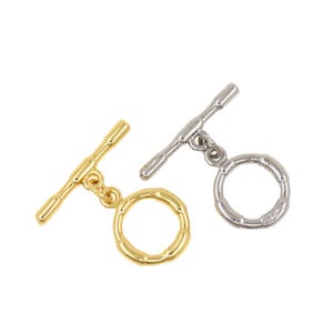 Pode incluir: Duas fechos de alternância, um dourado e outro prateado, são exibidos sobre um fundo branco. Cada fecho apresenta uma barra e um anel circular com um design texturizado. Estes fechos são comumente usados na fabricação de joias.