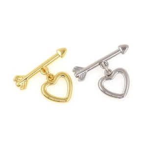 Puede incluir: Dos colgantes metálicos, uno dorado y otro plateado, cada uno con una flecha y un colgante de corazón. La flecha es horizontal, con el corazón colgando debajo. El corazón es un contorno abierto. El fondo es blanco.