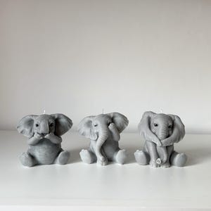 Può includere: Tre candele a forma di elefante grigio, ciascuna in una posa diversa: una si copre gli occhi, un'altra si copre le orecchie e l'ultima si copre la bocca. Le candele sono disposte in fila su una superficie bianca.