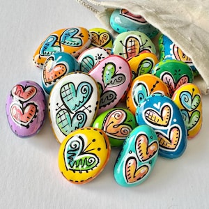 May include: A collection of painted heart-shaped rocks in various colors, including pink, blue, green, yellow, and orange. The rocks are decorated with swirls, dots, and lines.