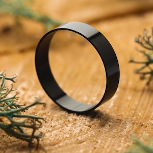 May include: A black ring with a smooth, polished finish. The ring is circular and rests on a textured, light brown surface, with sprigs of green foliage in the background. The ring's design is simple and modern.