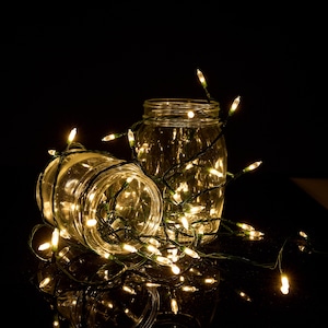 May include: Two clear glass jars illuminated by a string of warm white fairy lights. The green wire of the light strand is wrapped around and inside the jars, creating a glowing effect against the dark background with bright light points.