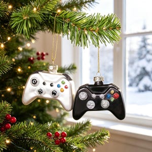 May include: Two video game controller ornaments hanging from a Christmas tree. One ornament is glossy white with silver glitter accents on the buttons and bumpers, while the other is glossy black with silver, blue, green, and red glitter details.
