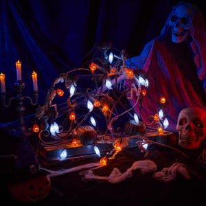 May include: A string of Halloween lights featuring small, glowing orange pumpkins and white ghost-shaped bulbs. The lights are draped over a dark, ornate metal structure, creating a festive display with a spooky aesthetic for seasonal decor.
