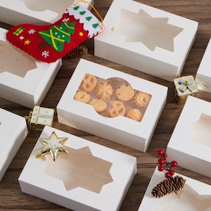 May include: A collection of white rectangular cardboard boxes with clear windows, some featuring star-shaped cutouts. One box contains assorted butter cookies. Decorations include a red felt Christmas stocking, gold gift ornaments, and a pinecone.