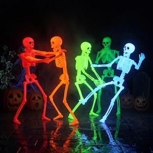 May include: Four articulated plastic skeletons in vibrant neon colors, including red, orange, green, and blue, posed in dynamic positions. The skeletons are illuminated against a dark background, with carved jack-o'-lanterns visible at their feet.