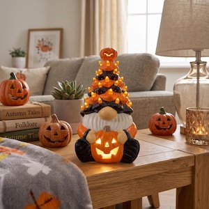 May include: A ceramic gnome figurine featuring a tall, conical hat decorated with small orange lights, a white beard, and black boots. The gnome holds a glowing jack-o'-lantern, while a small pumpkin sits atop the hat, creating a festive display.