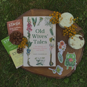 Puede incluir: Un libro titulado "Old Wives' Tales: The History of Remedies, Charms and Spells" de Mary Chamberlain, con una ilustraci&oacute;n floral en la portada. El libro est&aacute; rodeado de bolsitas de t&eacute;, peque&ntilde;os jabones redondos y pegatinas decorativas con dise&ntilde;os florales y de gatos.