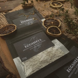 May include: A small, black pouch with a white zipper contains crushed eggshells. The pouch has a black label with white text that reads "EGGSHELL PROTECTION, CIRCLE CASTING, MANIFESTATION" and "10g".