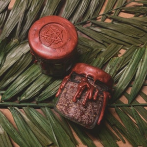 May include: Two small glass jars with brown lids. The jars are filled with a brown, grainy substance. One jar has a pentagram design on the lid.