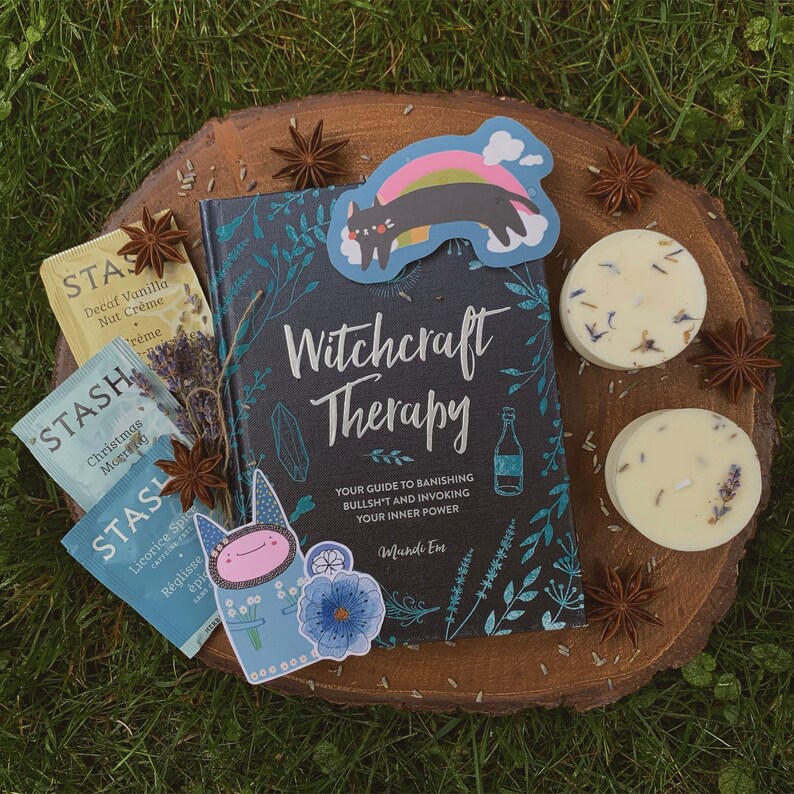Puede incluir: Un libro negro con el t&iacute;tulo "Witchcraft Therapy: Your Guide to Banishing Bullsh*t and Invoking Your Inner Power" de Mandi Em. El libro est&aacute; rodeado de bolsitas de t&eacute;, velas y pegatinas decorativas.