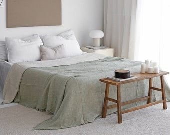 May include: A bedroom scene featuring a bed with white pillows and a sage green blanket. A wooden bench holds candles and a decorative box. A neutral-toned artwork hangs above the bed, and a white lamp sits on a bedside table.