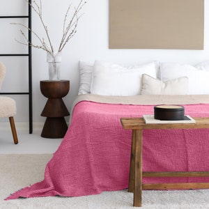 May include: A pink cotton throw blanket with a textured waffle weave pattern. The blanket is draped over a bed with white pillows and a wooden bench.