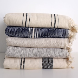 Può includere: Una pila di cinque coperte di cotone piegate in vari colori. Le coperte sono color crema, blu navy e grigio, con motivi a righe e a spina di pesce. Ogni coperta ha bordi sfrangiati e la pila è su una superficie bianca.